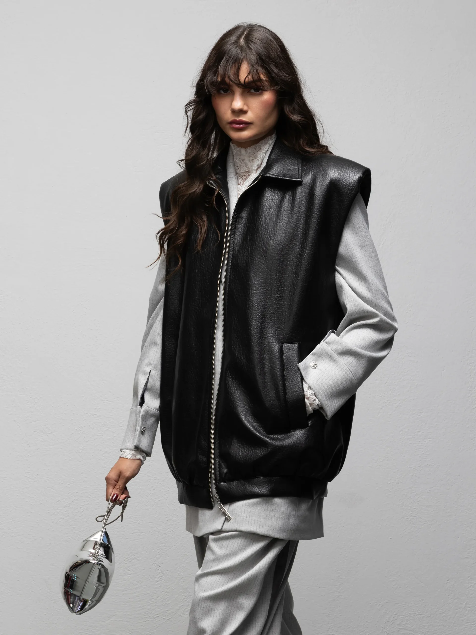 Black Vinyl gilet by We Are, close-up front view styled over a grey shirt with one hand in the side pocket